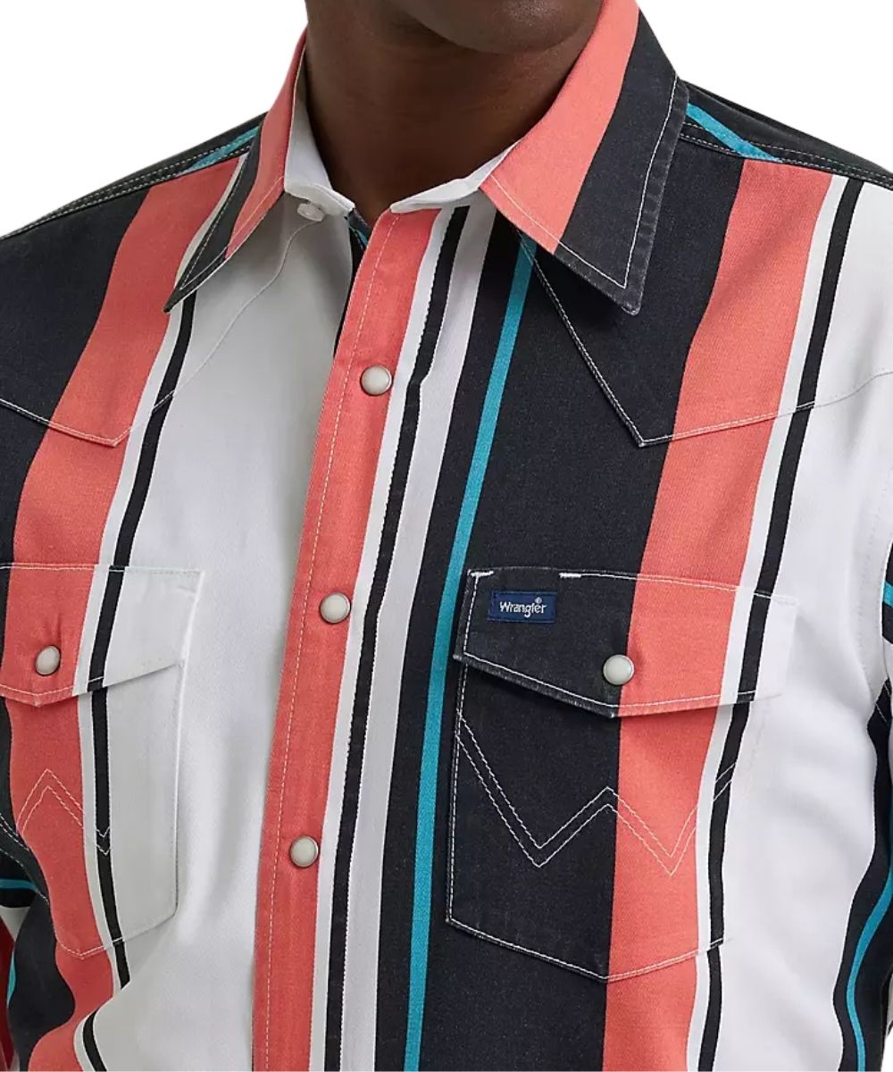 Close-up of a striped western-style button-up shirt on a model, featuring vertical stripes in coral pink, dark navy, white, and turquoise, with a visible Wrangler brand patch on the chest pocket.