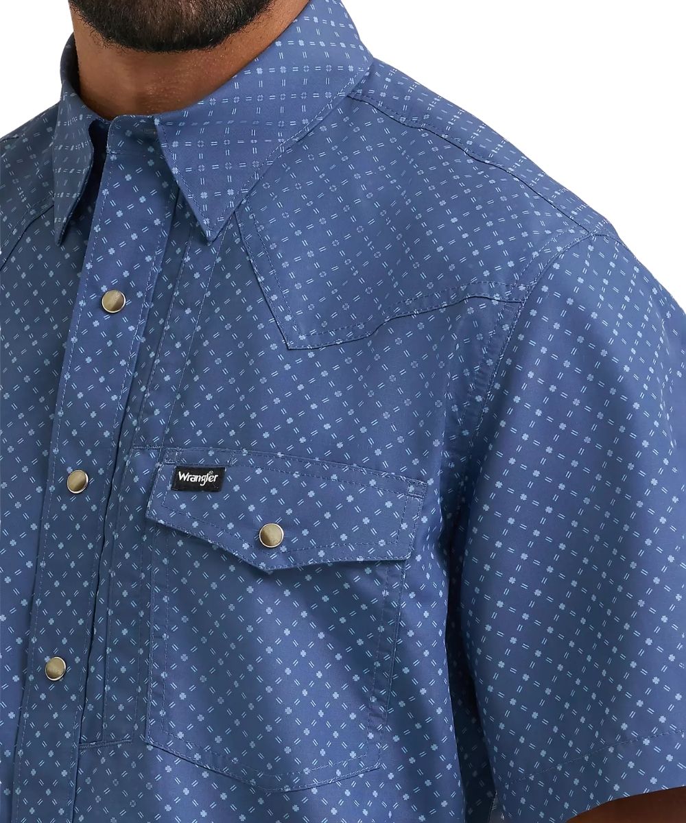 Close-up of a man wearing a Wrangler short-sleeve western shirt in blue with a small white geometric pattern, featuring a chest pocket and snap-button closure.
