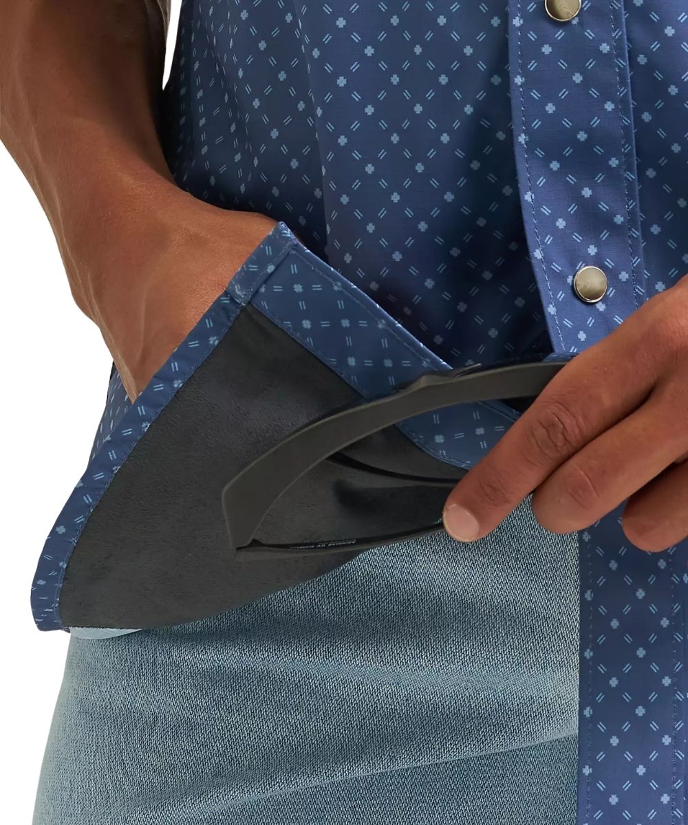 Close-up of a person wearing a blue patterned button-up shirt and light blue denim shorts, with one hand pulling a black curved strap out of a pocket.