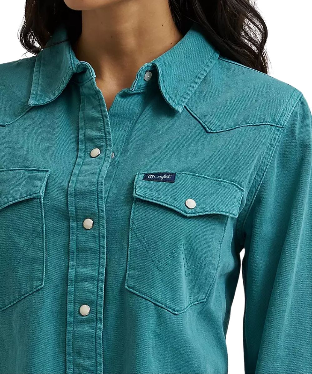 A close-up of a woman wearing a teal western-style button-up shirt with two chest pockets and a visible Wrangler label on the left chest pocket.