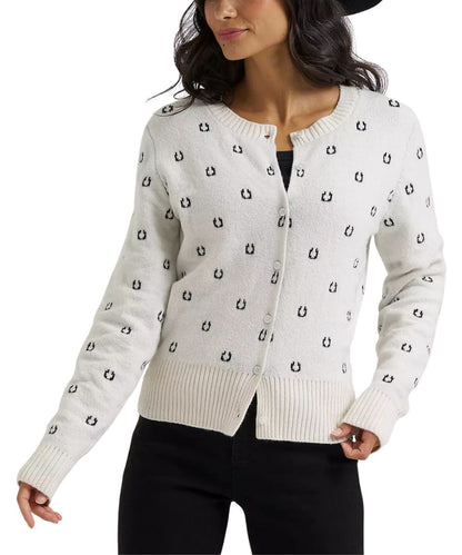 A woman wearing a cream-colored knit cardigan with a black horseshoe pattern, paired with black pants and a black hat, against a plain white background.