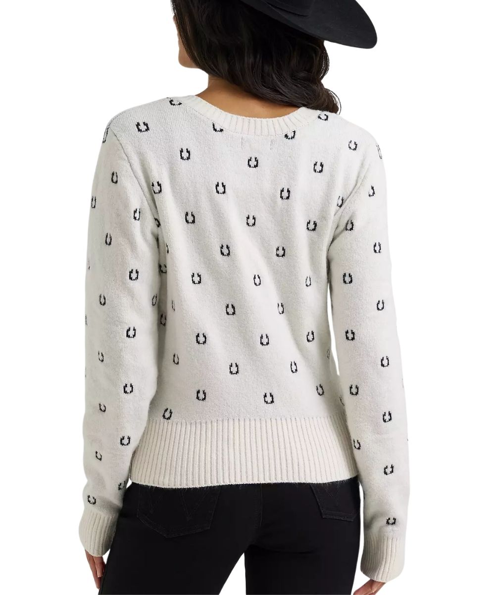 A woman shown from the back wearing a cream-colored knit sweater with a repeating black embroidered horseshoe pattern, paired with black pants and a black hat against a white background.