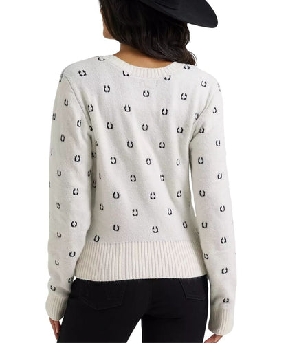 A woman shown from the back wearing a cream-colored knit sweater with a repeating black embroidered horseshoe pattern, paired with black pants and a black hat against a white background.
