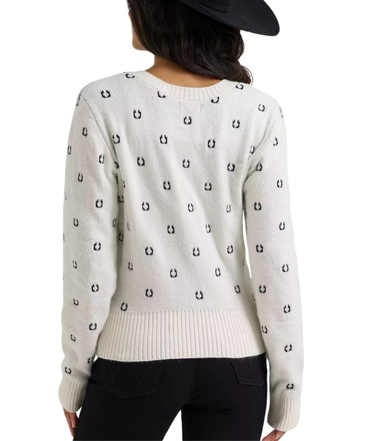 A woman shown from the back wearing a cream-colored knit sweater with a repeating black embroidered horseshoe pattern, paired with black pants and a black hat against a white background.