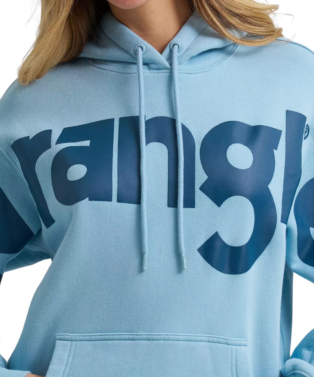 A close-up of a person wearing a light blue hoodie with a large dark blue 'wrangler' logo across the chest, featuring drawstrings and a front kangaroo pocket.