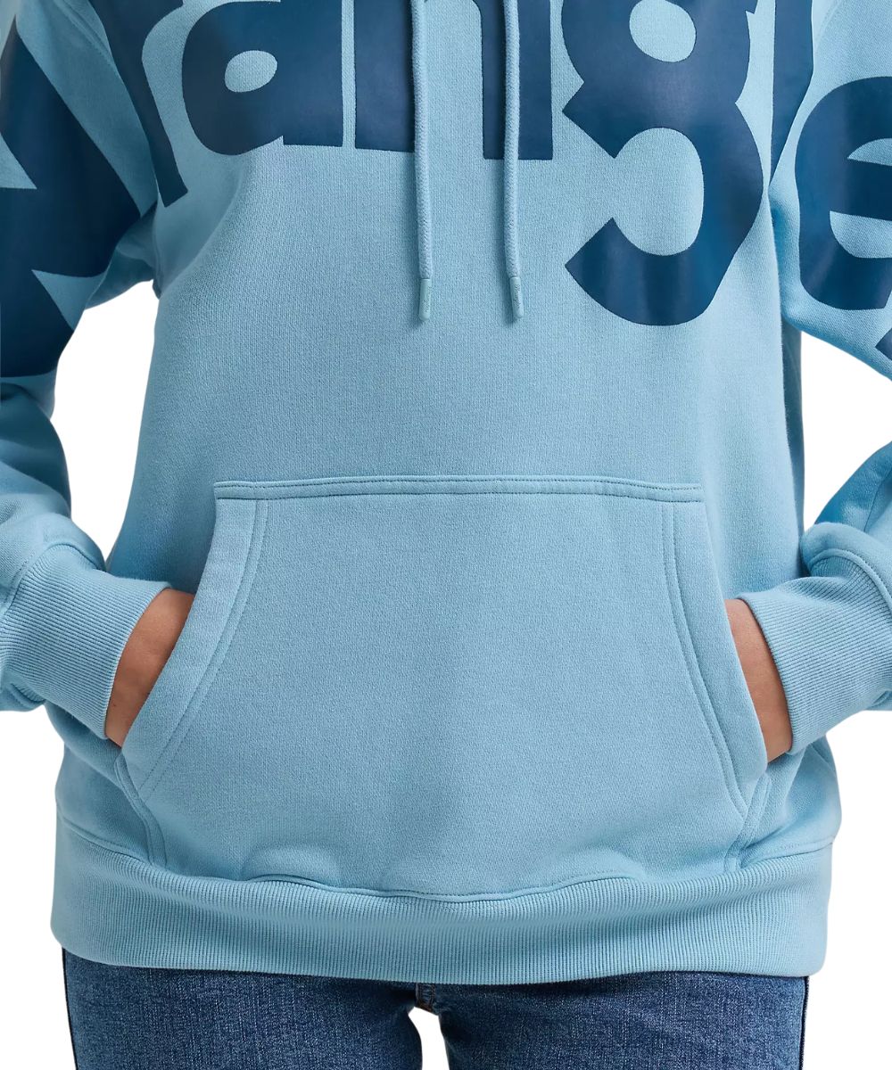 A close-up of a person wearing a light blue pullover hoodie with a large dark blue 'Wrangler' logo across the chest and matching drawstrings. The person's hands are in the front kangaroo pocket, and blue jeans are visible at the bottom against a plain white background.