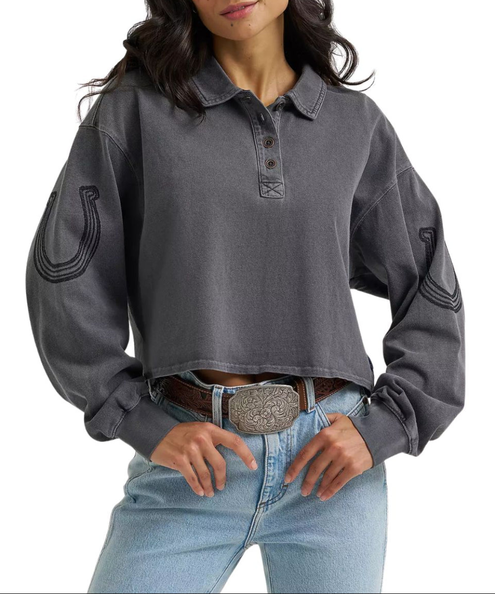 A woman wearing a dark gray cropped long-sleeve polo shirt with embroidered horseshoe details on the sleeves, paired with light blue jeans and a brown belt.