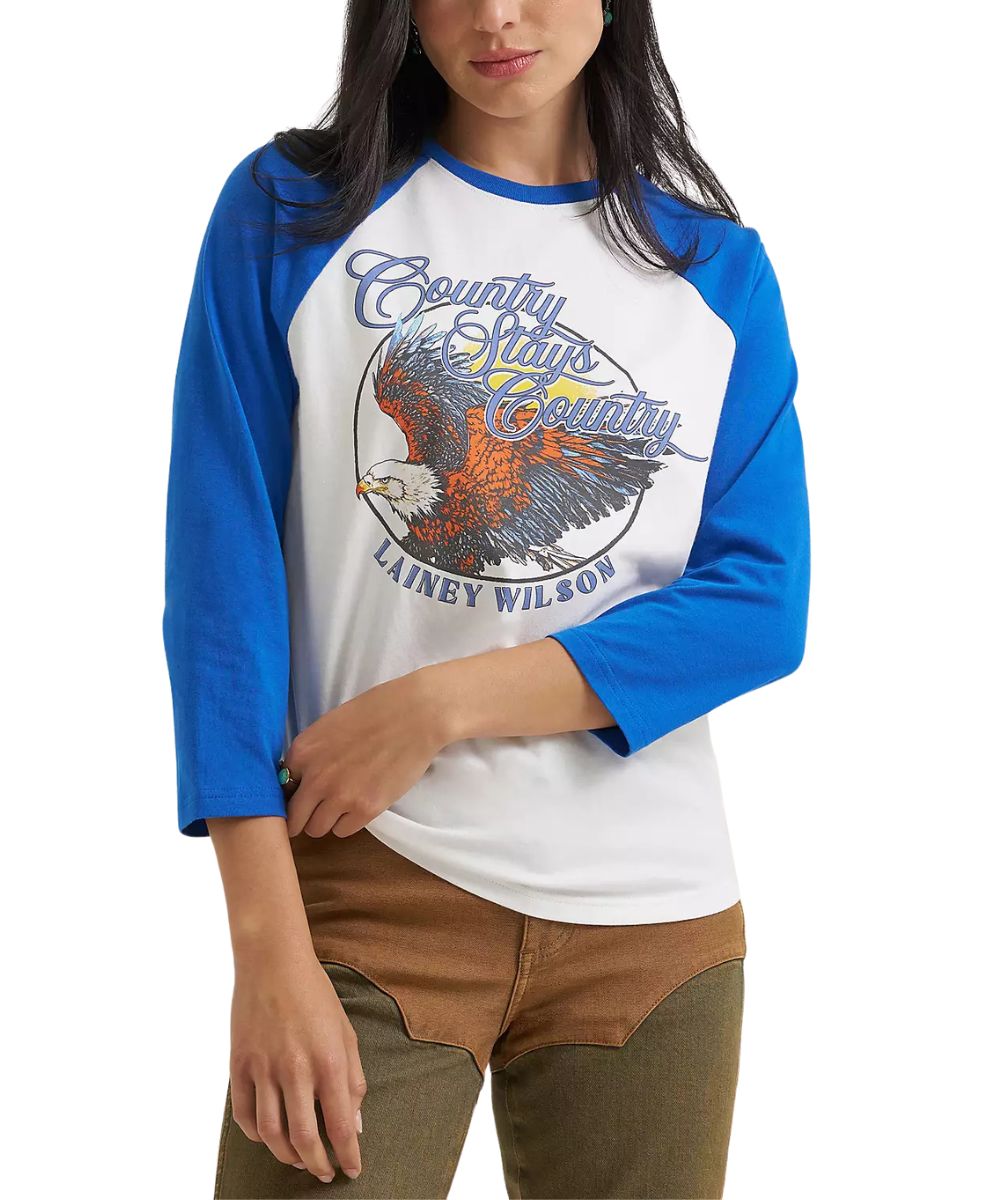A woman wearing a 3/4-sleeve raglan baseball t-shirt with a white body, blue sleeves, and a large graphic on the front featuring an eagle and the text 'Country Stays Country' and 'Lainey Wilson'.
