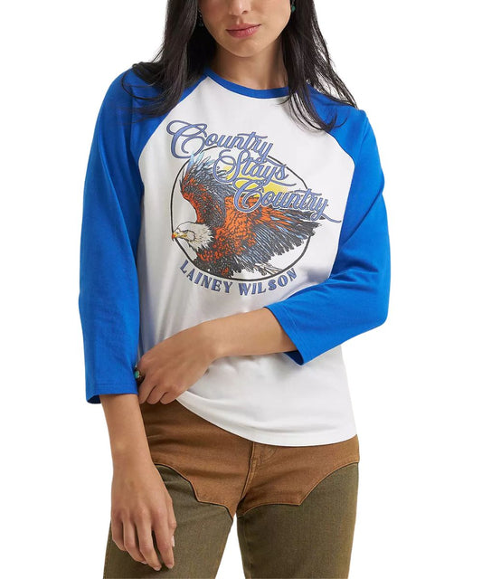 A woman wearing a 3/4-sleeve raglan baseball t-shirt with a white body, blue sleeves, and a large graphic on the front featuring an eagle and the text 'Country Stays Country' and 'Lainey Wilson'.