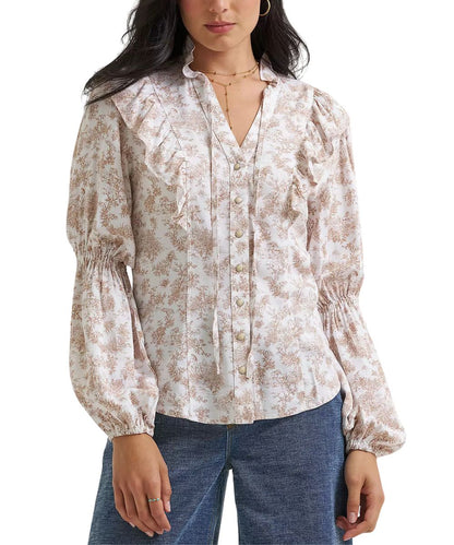 A woman wearing a long-sleeve button-front blouse with a beige and brown floral toile pattern, ruffled collar details, and gathered elastic cuffs, paired with blue jeans against a plain white background.