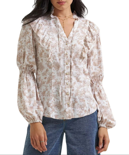 A woman wearing a long-sleeve snap-front blouse with a beige and brown floral toile pattern, ruffled collar details, and gathered elastic cuffs, and a v-neckline with self-tie closure