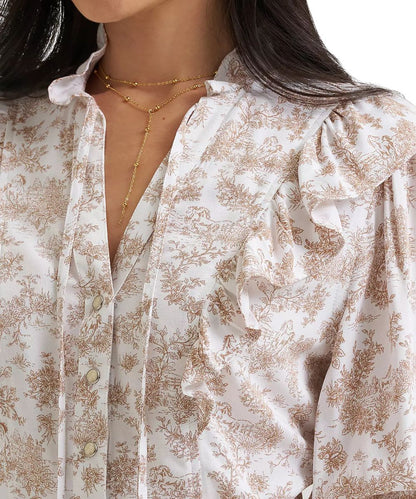 A close-up of a woman wearing a white satin blouse with a brown toile floral pattern, featuring ruffled details on the shoulders and a gold Y-necklace.