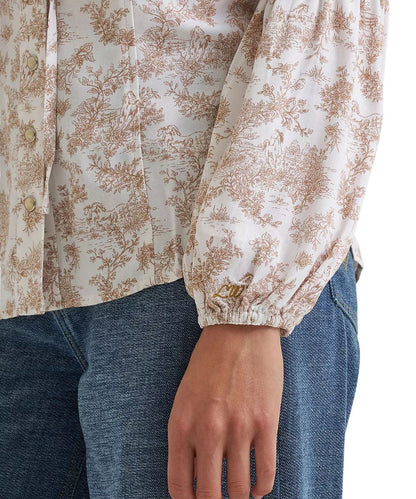 A close-up of a person wearing a cream-colored blouse with a brown toile floral pattern and blue jeans. The blouse features gathered, balloon-style sleeves with elastic cuffs, and a button-down front.