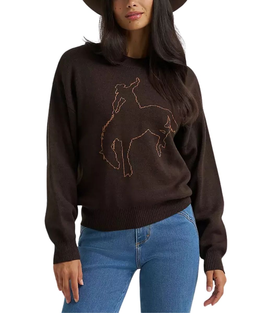 A woman wearing a dark brown knit sweater with a light brown outline graphic of a bucking horse on the front, paired with blue jeans and a dark wide-brim hat, against a plain white background.