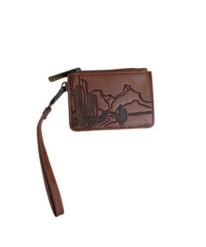 A brown leather zippered wristlet wallet featuring an embossed desert landscape design with cacti and mountains, complete with a matching wrist strap, shown on a plain white background.