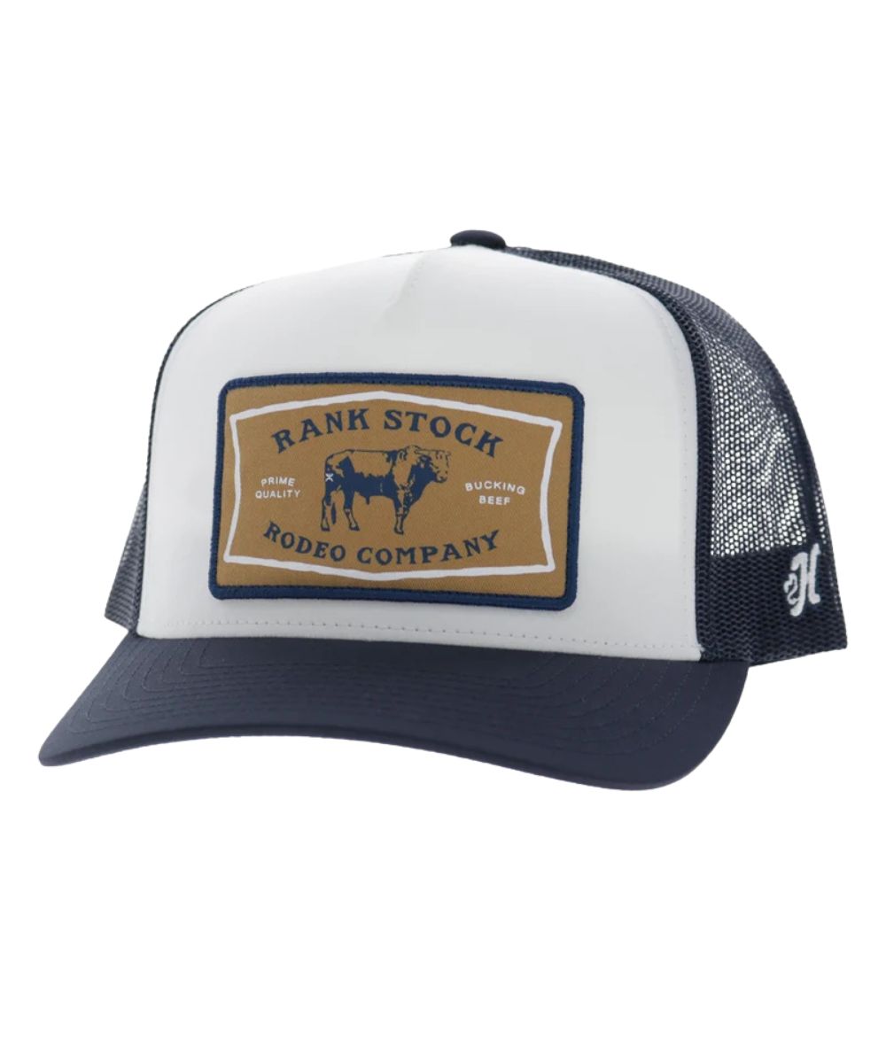 Left angled view of the unisex adult's Hooey Rank Stock Trucker Snapback Cap featuring the white front with a tan rectangle patch that reads: "Rank Stock Rodeo Company - High Quality - Bucking Beef" with a bull graphic, navy curved bill, and navy mesh sides