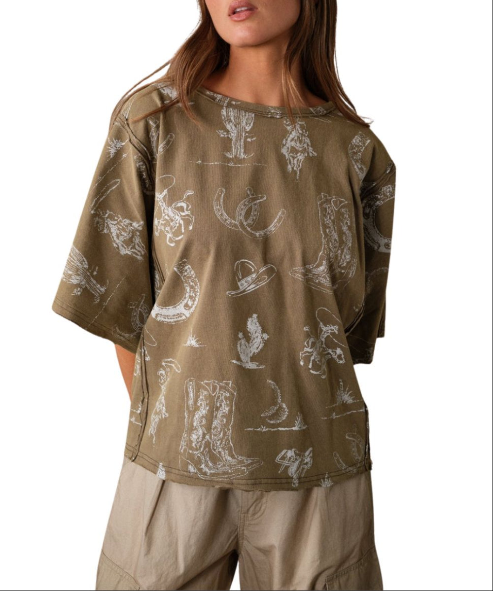 A woman wearing an oversized short-sleeve t-shirt with a distressed olive base and a white western-themed print featuring boots, hats, and horseshoes