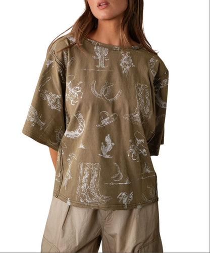 A woman wearing an oversized short-sleeve t-shirt with a distressed olive base and a white western-themed print featuring boots, hats, and horseshoes