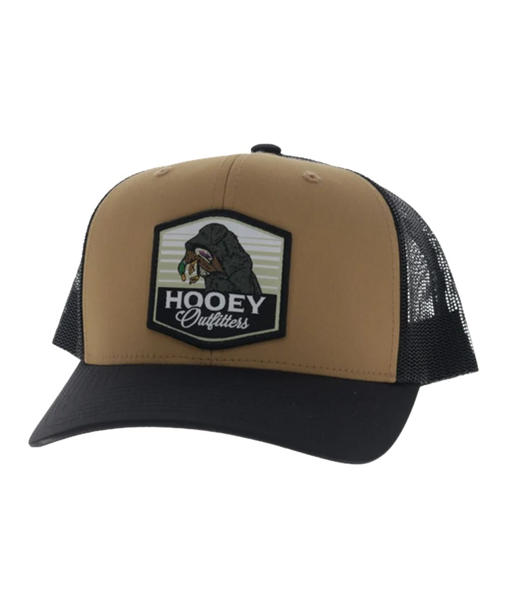 Left angled view of the unisex adult's Hooey Toller Trucker Cap featuring the tan front with the Hooey Outfitters Lab Dog Patch, black curved bill, and black mesh sides