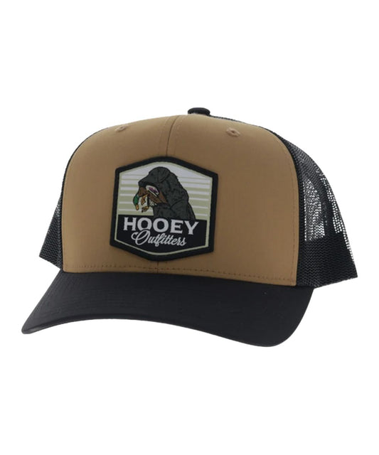 Left angled view of the unisex adult's Hooey Toller Trucker Cap featuring the tan front with the Hooey Outfitters Lab Dog Patch, black curved bill, and black mesh sides