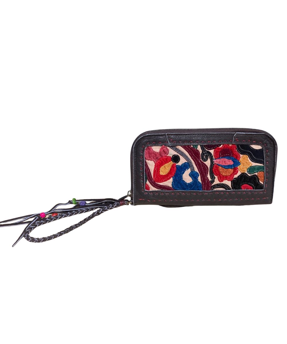 A dark brown leather zip-around wallet featuring a colorful embroidered front panel with red, blue, yellow, and black patterns, complete with a braided wrist strap.