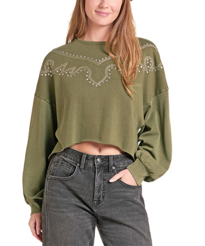 A woman wearing an olive green cropped long-sleeve sweatshirt with a western-style embroidered pattern across the chest, paired with dark gray jeans, against a plain white background.