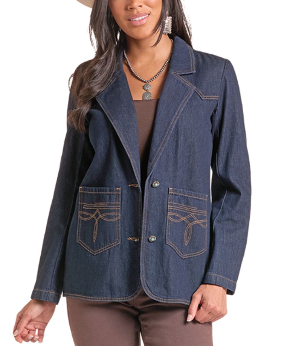 A woman wearing a dark blue denim blazer with contrast tan stitching, featuring two front patch pockets with decorative stitching, a brown top, and brown pants against a white background.