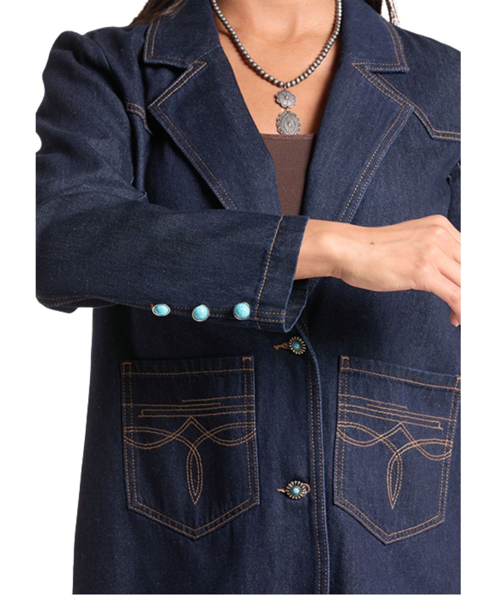A close-up of a person wearing a dark indigo denim blazer with contrast tan stitching, decorative turquoise-colored buttons on the cuff, and two large front patch pockets with stitched floral details.