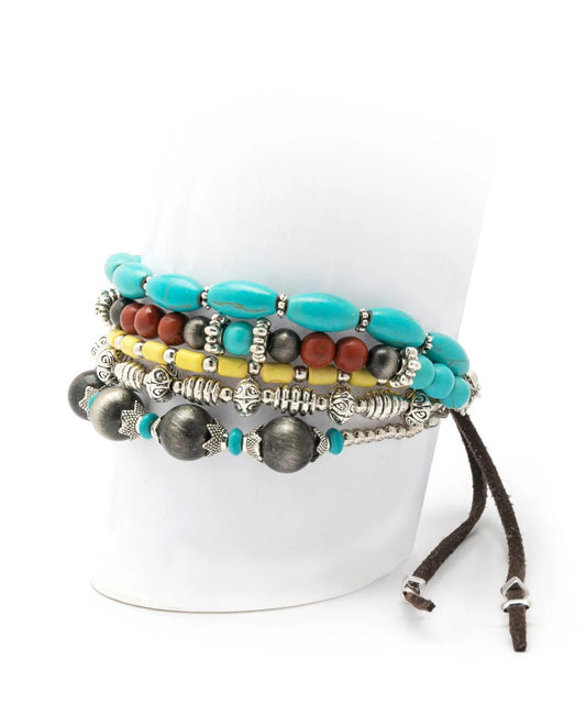 A stack of bohemian-style beaded bracelets displayed on a white cylindrical mannequin stand. The bracelets feature turquoise blue beads, dark gray textured beads, silver-toned metal spacers, a yellow beaded strand, and a brown cord with silver-toned end details.