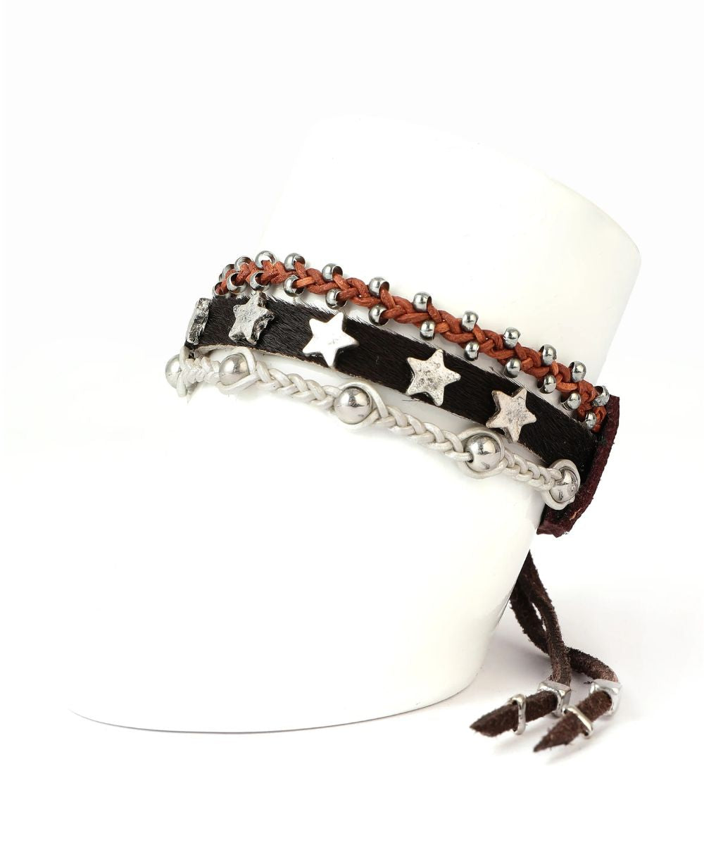 A set of bohemian-style multi-layered bracelets displayed on a white mannequin arm. The bracelets feature dark brown leather straps, a reddish-brown braided cord, silver-tone metal beads, star-shaped charms, braided chains, and dangling leather tassels.