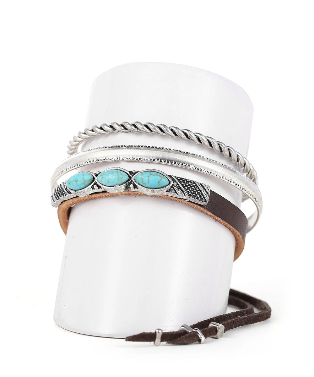 A stack of western-style bracelets displayed on a white cylindrical stand. The set includes silver-toned twisted rope, textured, and smooth bands, a brown leather band with three oval turquoise stones, and a dark brown suede wrap with silver hardware.
