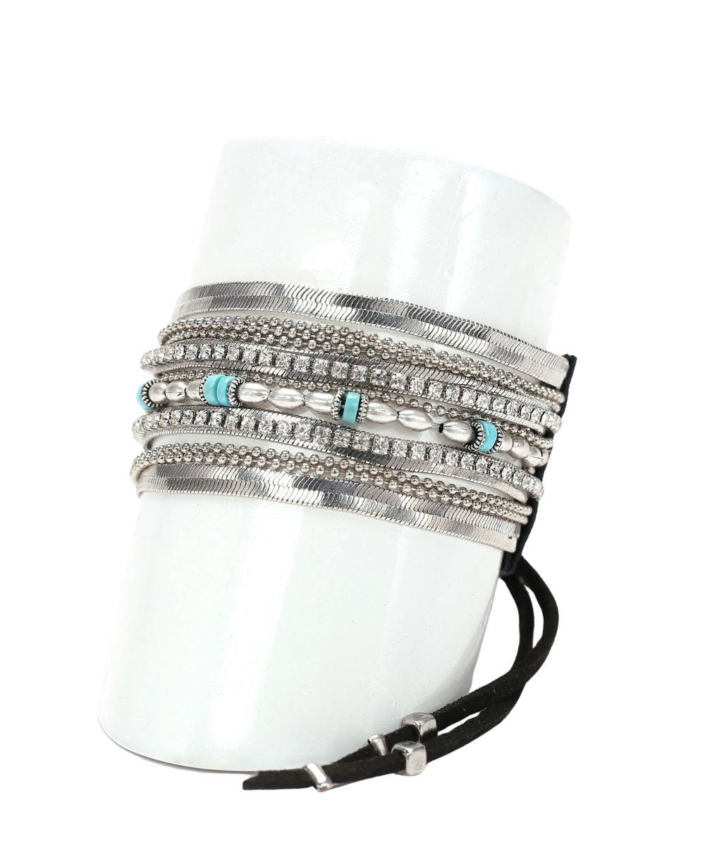 A multi-strand silver bracelet cuff with textured bands, rhinestone rows, turquoise bead accents, and a black wrap-around cord with silver hardware, displayed on a white cylindrical mannequin stand.
