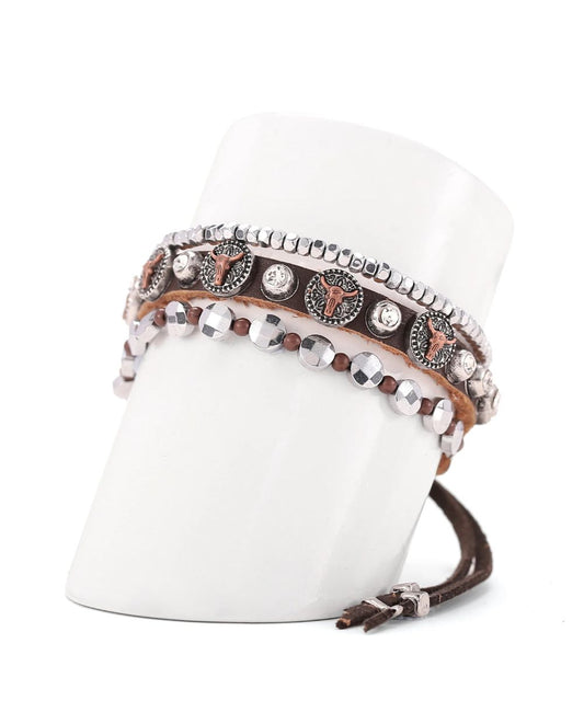 A set of western-style wrap bracelets displayed on a white cylindrical stand. The bracelets feature brown leather, silver-tone studs, clear rhinestones, and circular silver-toned medallions with brown western motifs.