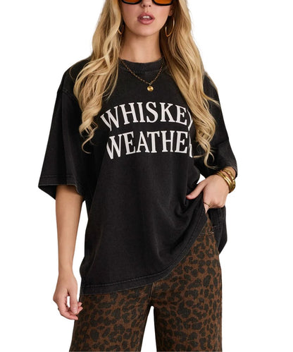 A woman wearing a black acid-wash graphic t-shirt with 'WHISKEY WEATHER' printed in white arched text, paired with brown leopard print pants, against a plain white background.