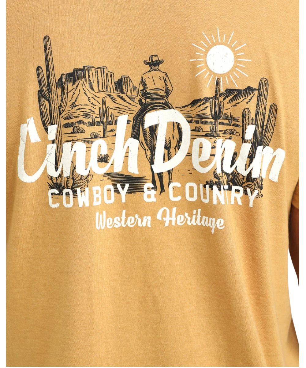 A close-up of a tan graphic t-shirt featuring a western theme with a cowboy, cacti, and a sun, with the text 'Cinch Denim' and 'Cowboy & Country Western Heritage' printed in white.