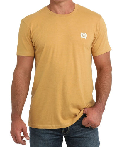 A man wearing a mustard yellow crewneck t-shirt with a small white chest graphic, paired with blue jeans, against a plain white background.