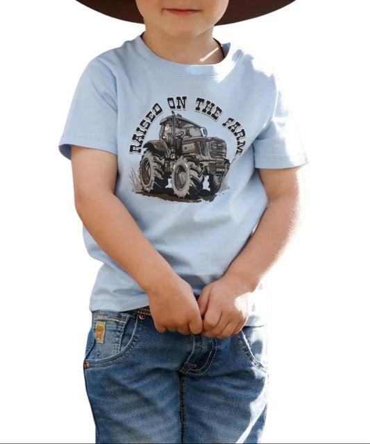 A toddler wearing a light blue short-sleeve t-shirt with a tractor graphic and the words 'RAISED ON THE FARM', paired with blue jeans and a dark brown cowboy hat, set against a plain white background.