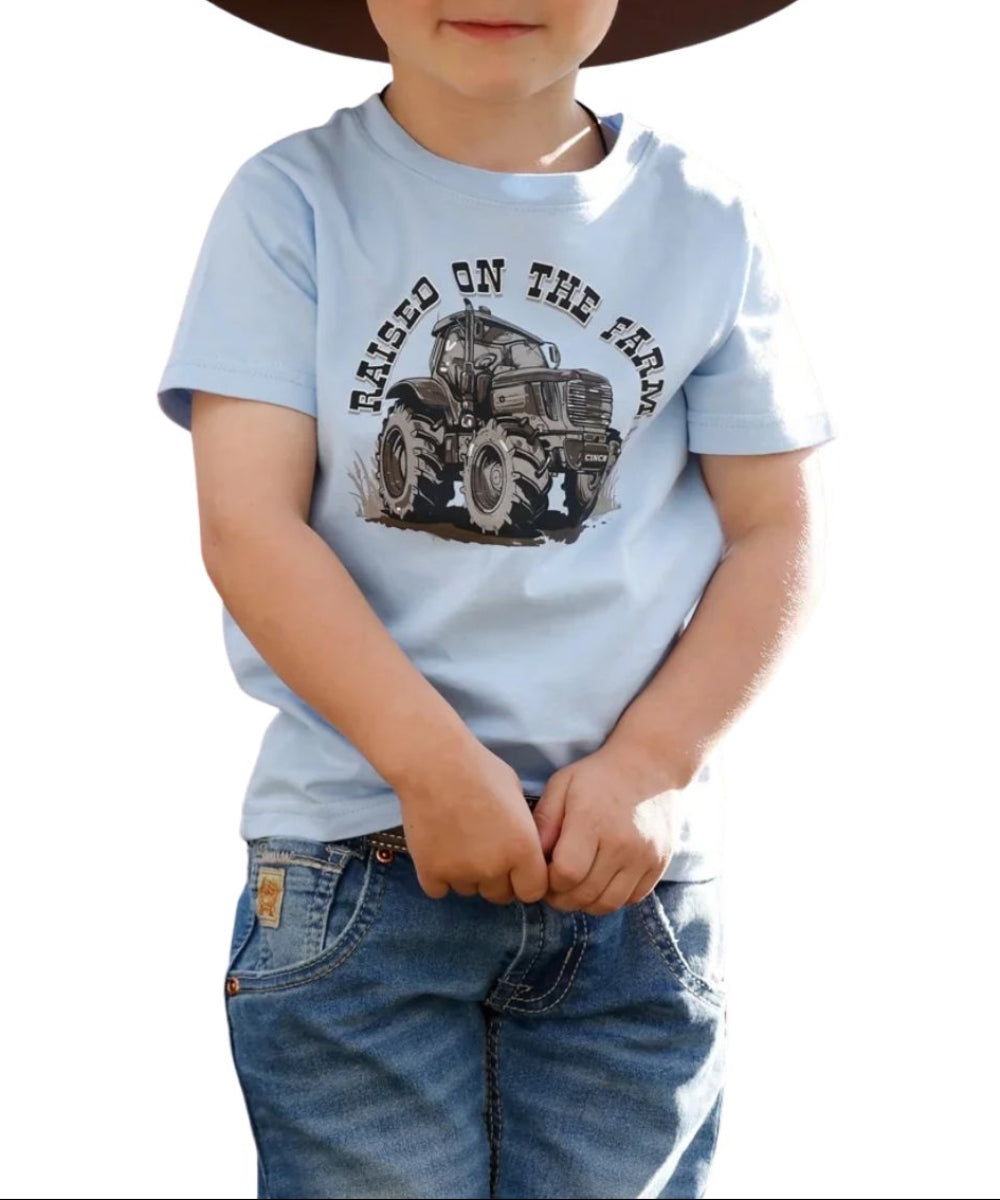 A toddler wearing a light blue short-sleeve t-shirt with a tractor graphic and the words 'RAISED ON THE FARM', paired with blue jeans and a dark brown cowboy hat, set against a plain white background.