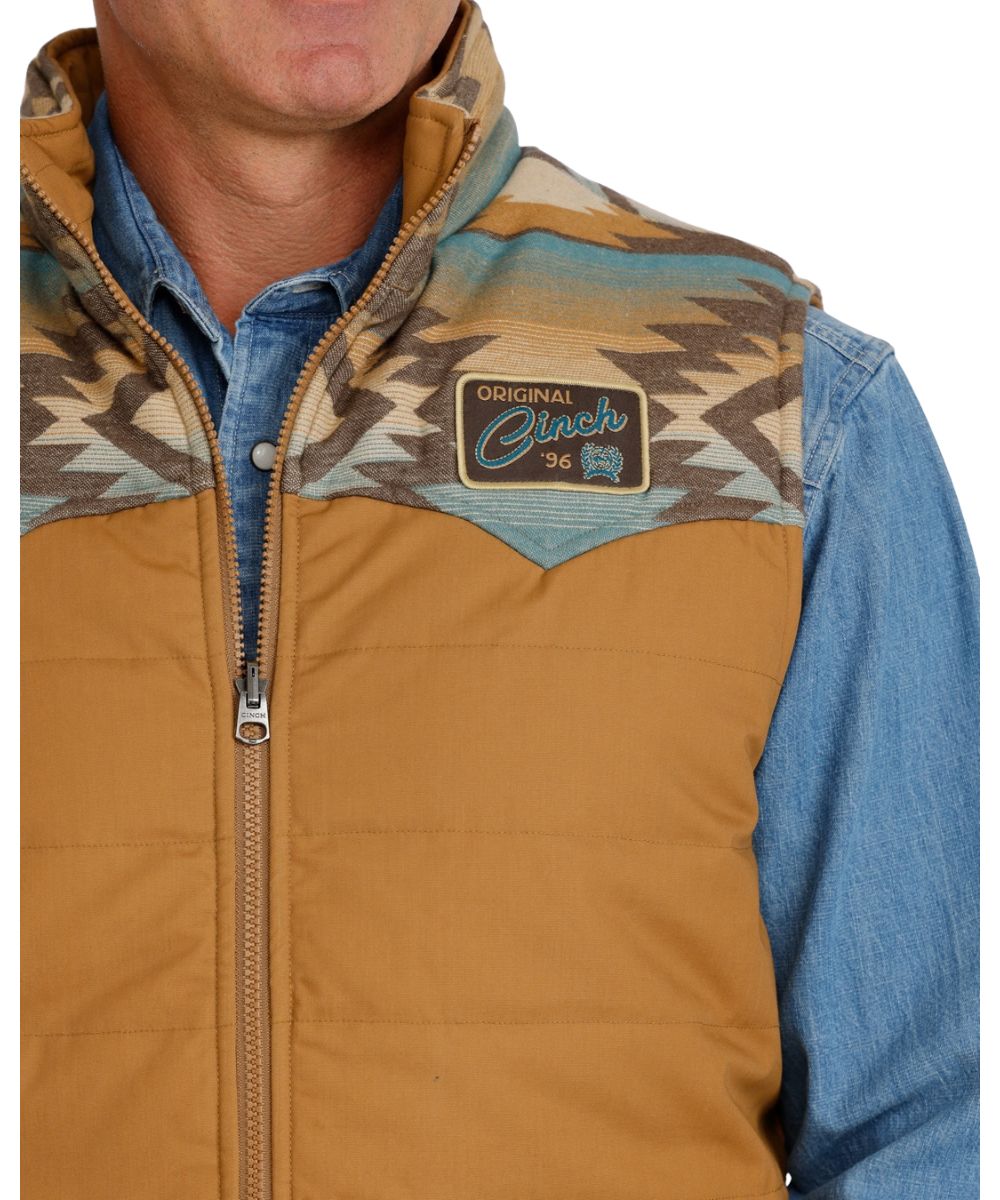Close-up view of the men's tan canvas vest featuring the woven logo patch at the left chest featuring the water-resistant finish and quilted design