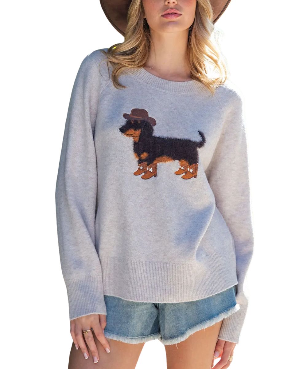 A woman wearing a light gray knit sweater with a brown and black dachshund graphic wearing a small brown hat, paired with denim shorts and a wide-brim hat against a white background.