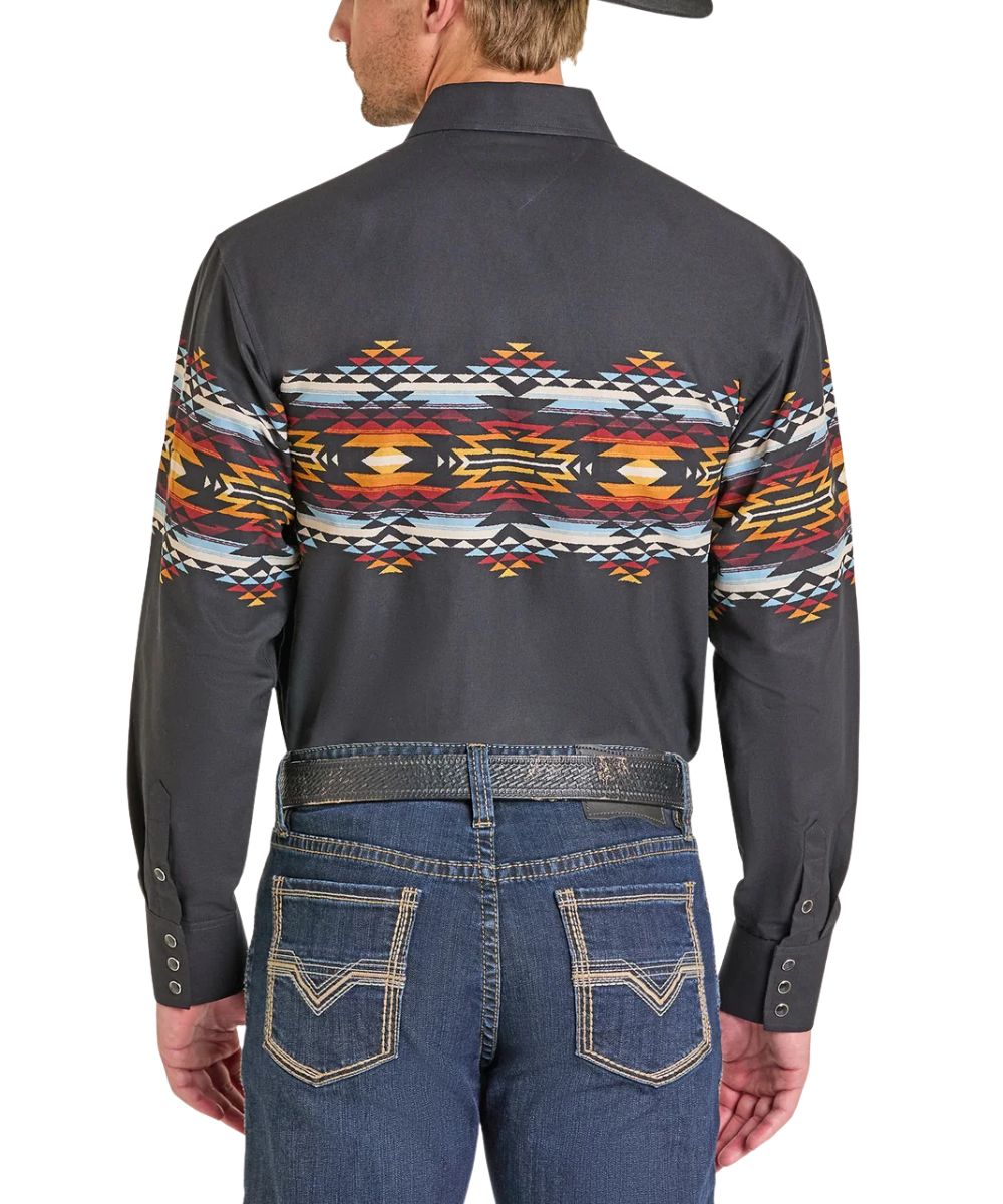 A man viewed from the back wearing a dark charcoal long-sleeve western shirt with a colorful geometric Southwestern pattern across the shoulders and upper back, paired with blue jeans and a dark belt.