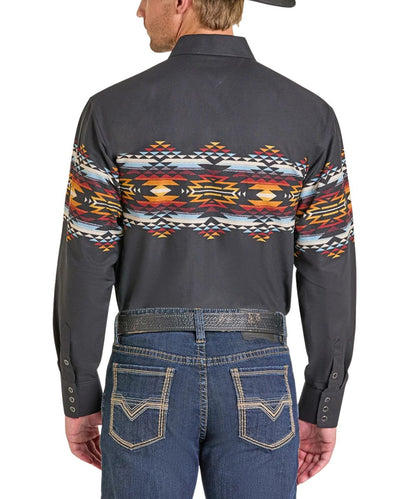 A man viewed from the back wearing a dark charcoal long-sleeve western shirt with a colorful geometric Southwestern pattern across the shoulders and upper back, paired with blue jeans and a dark belt.