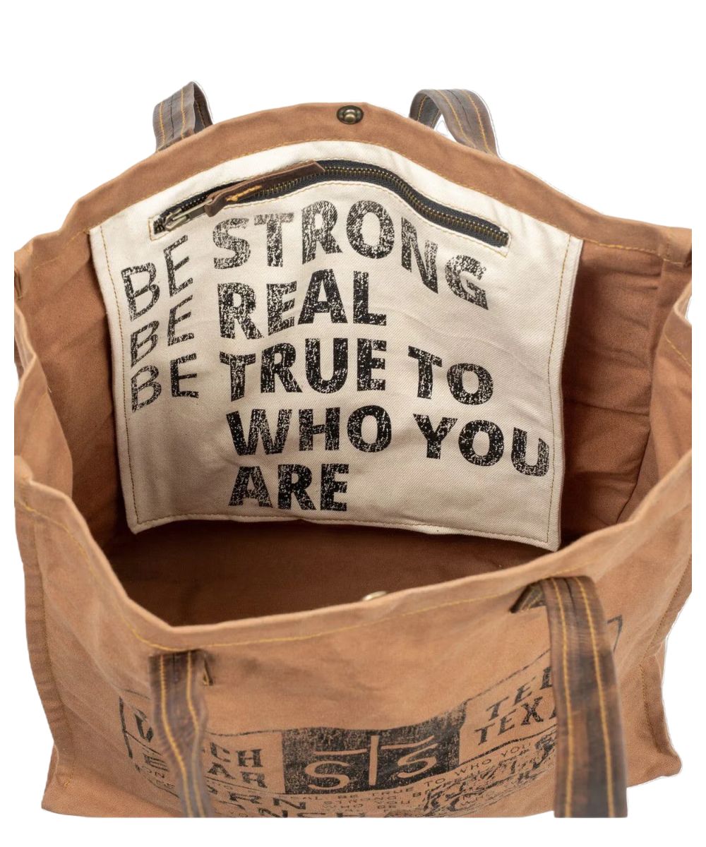 Interior view of the tote featuring the main compartment with a snap closure, an interior zip pocket, and an inside quote that reads: "Be Strong. Be Real. Be True To Who You Are."