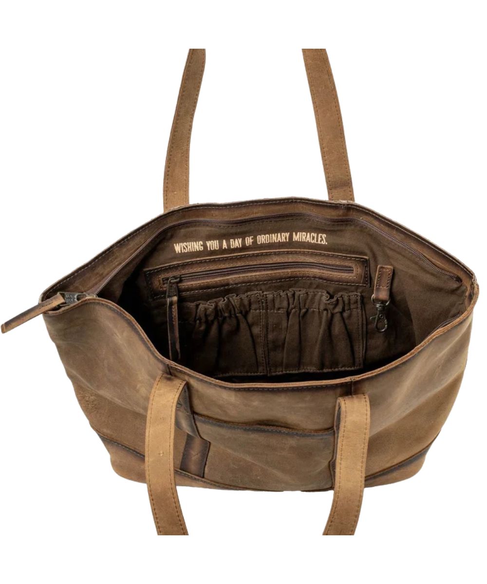 Interior view of the women's brown cowhide tote featuring the interior zip pocket, 2 interior scrunch pockets, an interior keychain clasp, and a quote above the zip pocket that reads: "Wishing You A Day Of Ordinary Miracles."