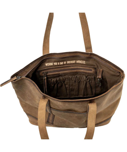 Interior view of the women's brown cowhide tote featuring the interior zip pocket, 2 interior scrunch pockets, an interior keychain clasp, and a quote above the zip pocket that reads: "Wishing You A Day Of Ordinary Miracles."