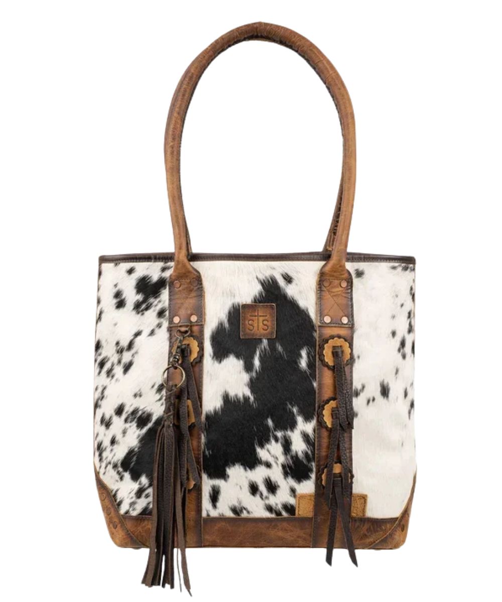 Front view of the women's brown STS Guadalupe Cowhide Palamino Tote Bag featuring the hair-on-hide front with a STS branded leather patch, dual rolled leather carry handles, front leather conchos with fringe accents, and a removable leather tassel