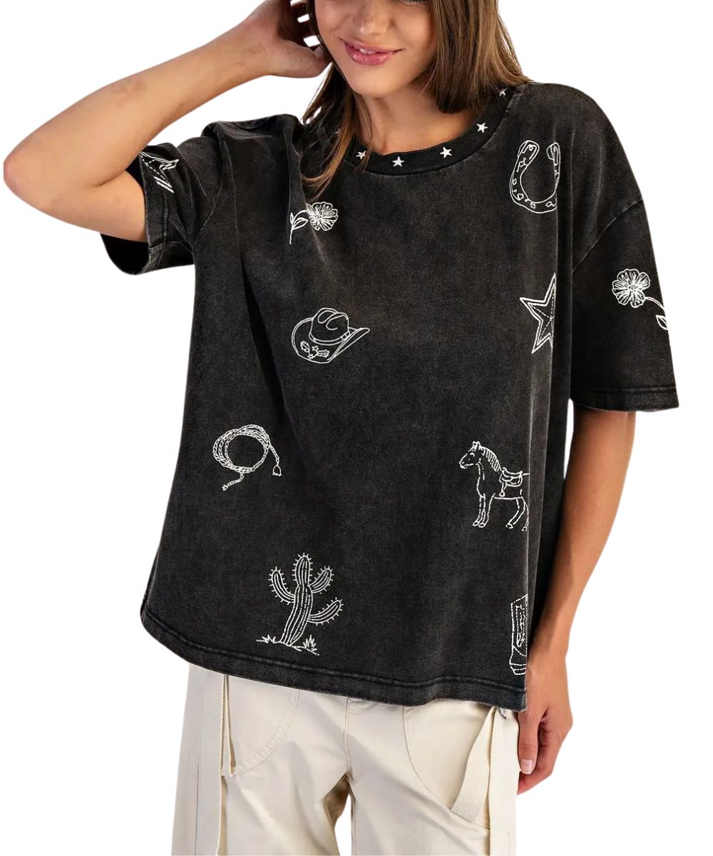 A woman wearing a black acid-wash t-shirt with white western-themed prints including stars, a cowboy hat, a lasso, a cactus, and a horse, paired with beige cargo pants against a white background.