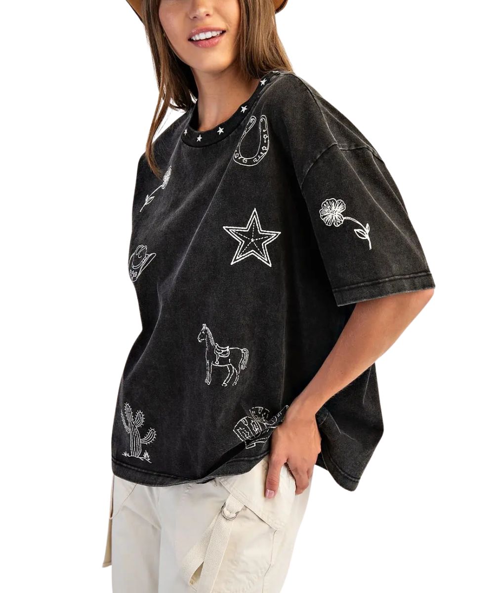 A woman wearing a faded black short-sleeve t-shirt with white embroidered western motifs including a star, horseshoe, cactus, and flower, paired with light beige cargo pants against a white background.
