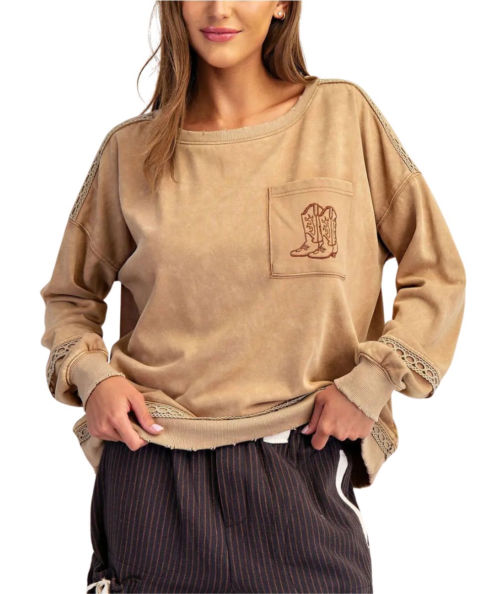 A woman wearing a brown long-sleeve western-style sweatshirt with a pocket featuring an embroidered cowboy boot design and lace trim on the sleeves, paired with dark pinstriped pants against a white background.