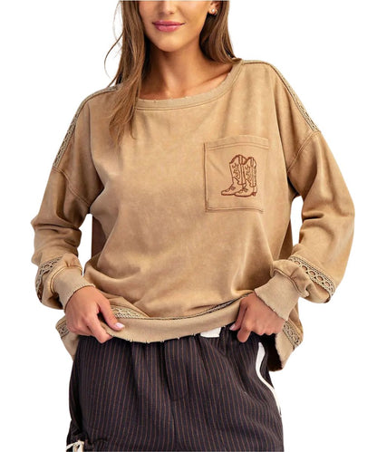 A woman wearing a brown long-sleeve western-style sweatshirt with a pocket featuring an embroidered cowboy boot design and lace trim on the sleeves, paired with dark pinstriped pants against a white background.